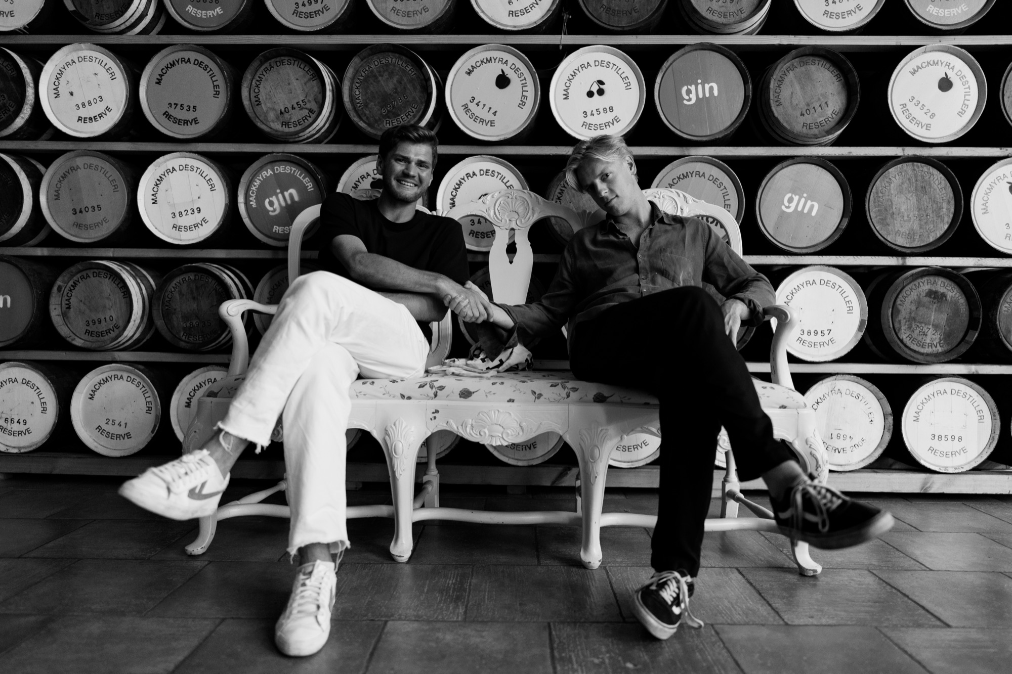 Black and white photo of two men shaking hands on an ornate bench in front of a wall of whisky barrels