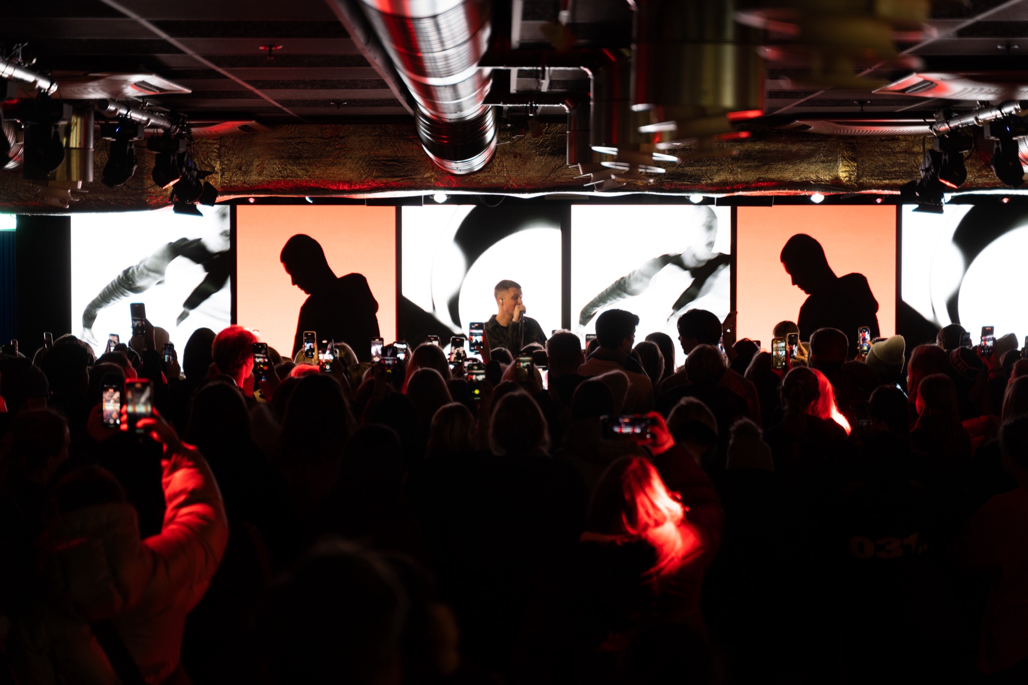 Artist performing on stage in front of large screens, crowd filming with their phones in red light