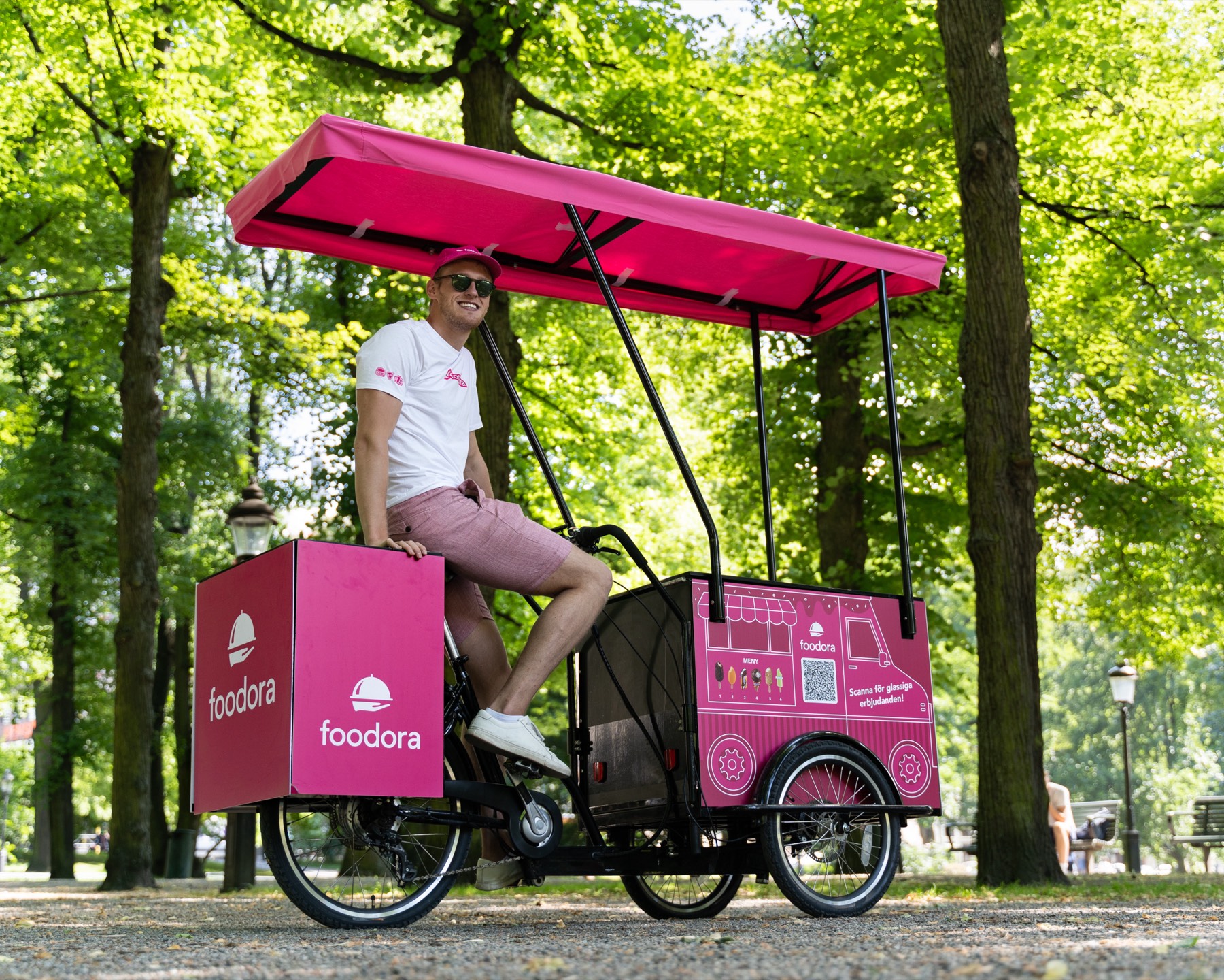 Smiling man on a pink foodora ice cream cargo bike with a canopy, in a leafy park